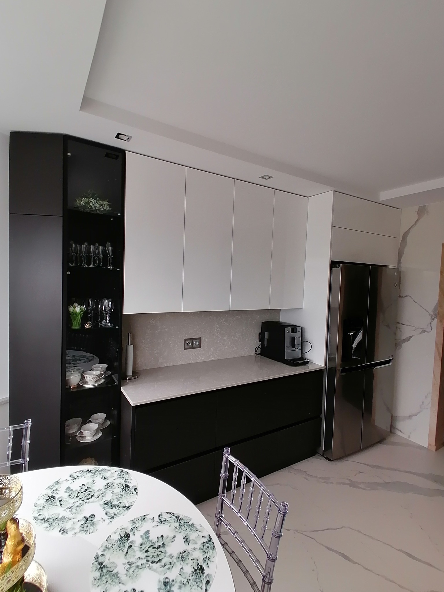 Nowoczesna kuchnia z białymi szafkami wiszącymi i czarnymi szafkami dolnymi, szklana witryna z dekoracyjnymi przedmiotami i okrągły stół z nakryciem w tle, jasna podłoga imitująca marmur.
