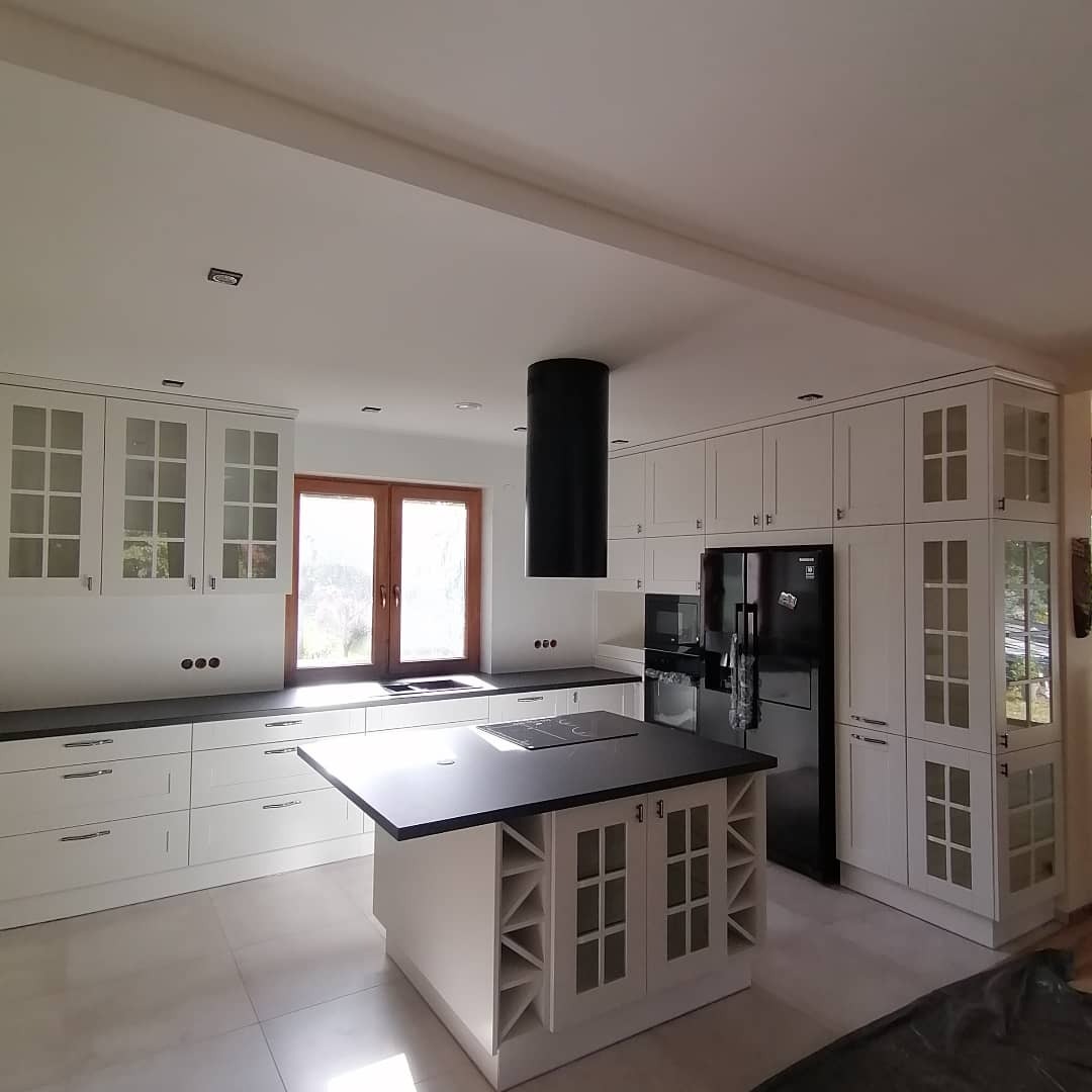 Jasna kuchnia z białymi szafkami z przeszklonymi frontami, czarnym blatem i wyspą kuchenną z miejscem na wino, okap cylindryczny w kolorze czarnym.