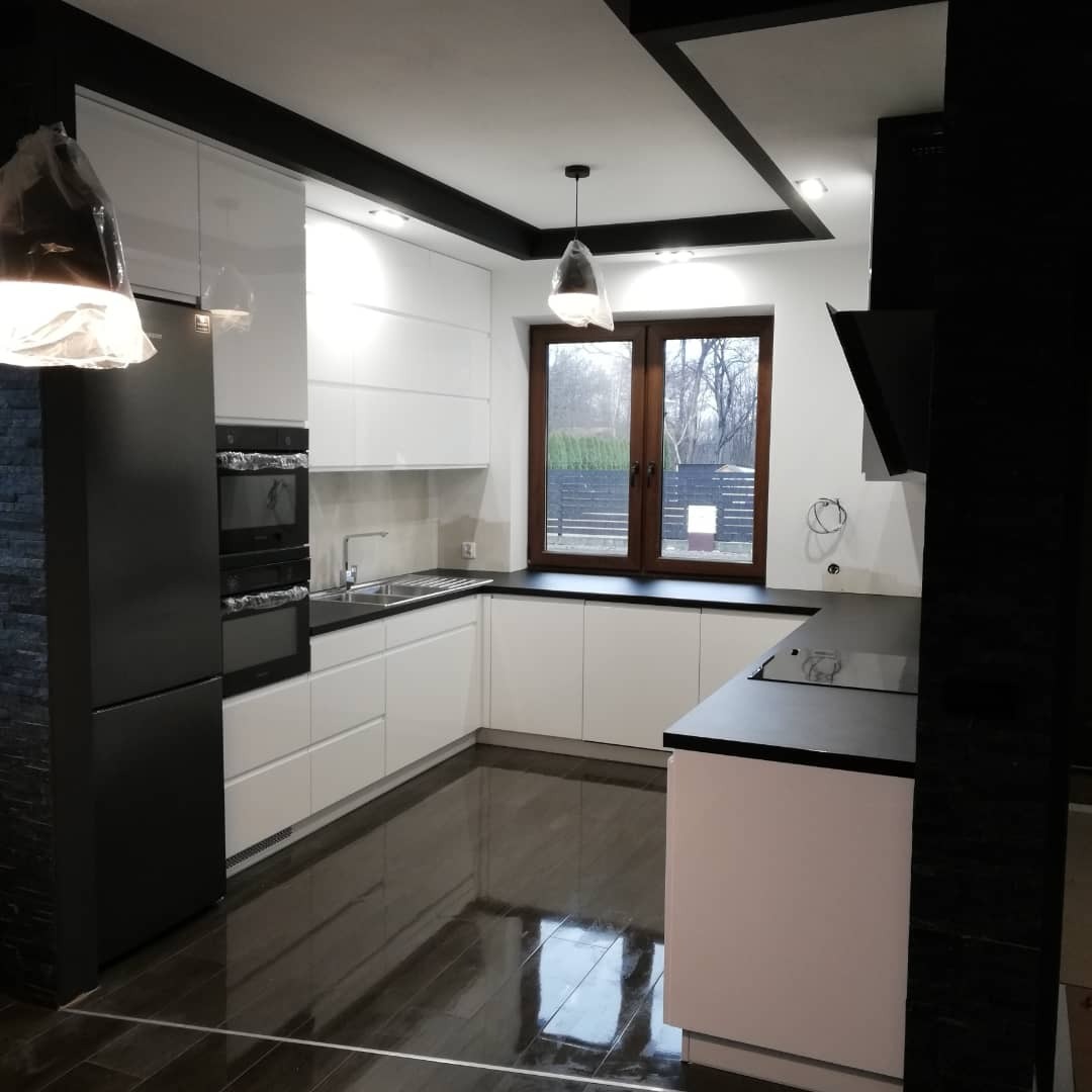 Nowoczesna kuchnia w bieli i czerni z błyszczącą podłogą, widok na okno z drewnianą ramą, lampy wiszące jeszcze w folii ochronnej.