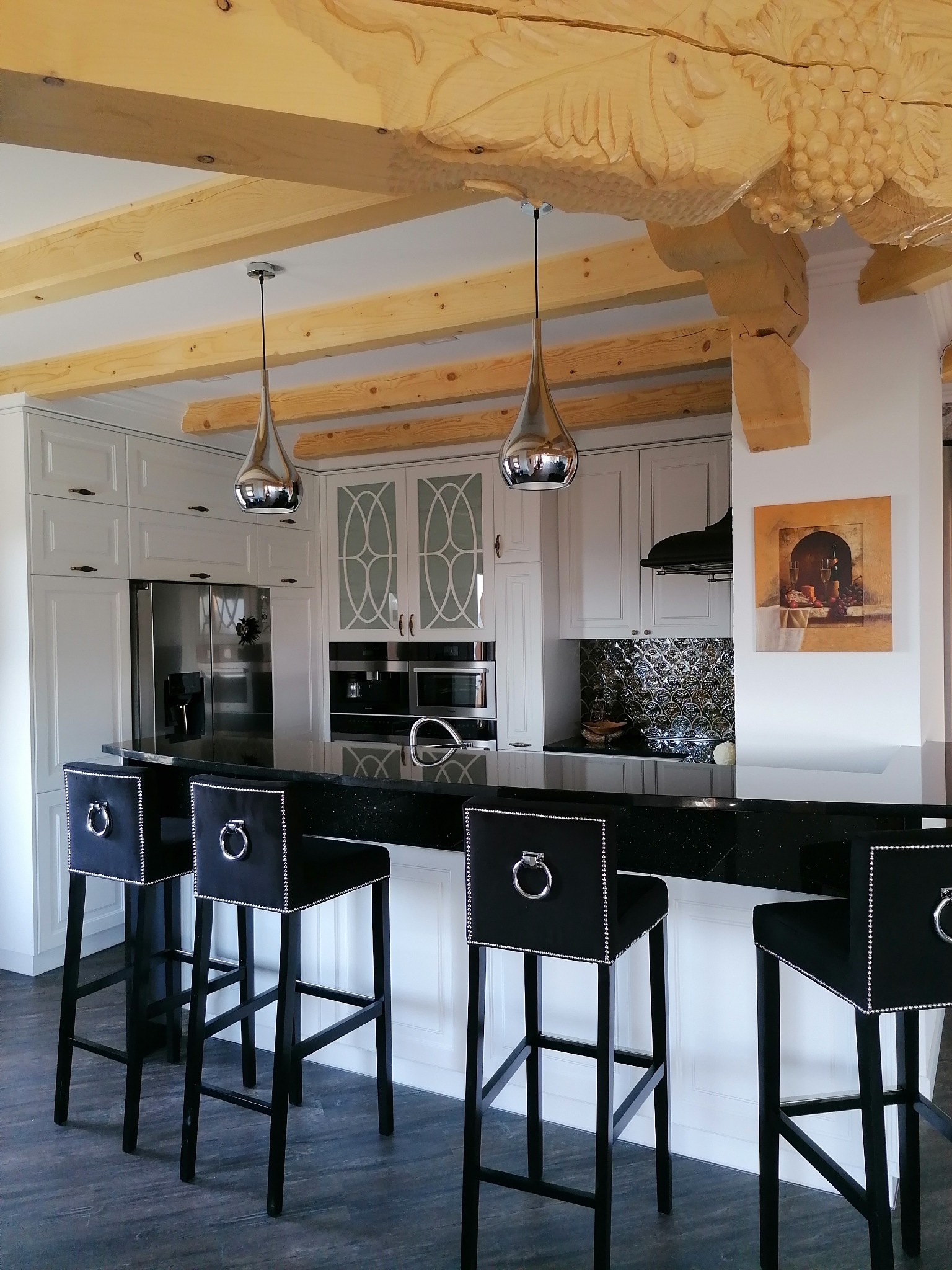 Nowoczesna kuchnia z białymi szafkami, czarnym blatem i hokerami z czarnej tkaniny z ozdobnymi srebrnymi nitami, drewniane belki stropowe z rzeźbieniami winogron.