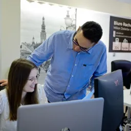 Pracownicy biura rachunkowego analizują dokumenty przy komputerze; w tle widoczny plakat reklamowy biura oraz zdjęcie panoramy miasta.