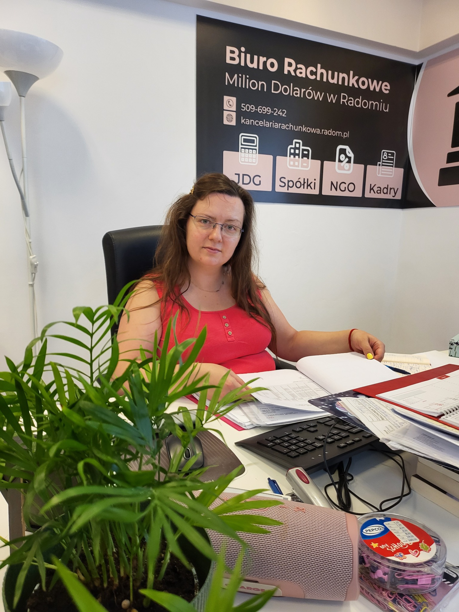 Kobieta w okularach siedzi za biurkiem w biurze rachunkowym, otoczona dokumentami i biurowymi akcesoriami, z rośliną doniczkową w pierwszym planie i szyldem firmy w tle.