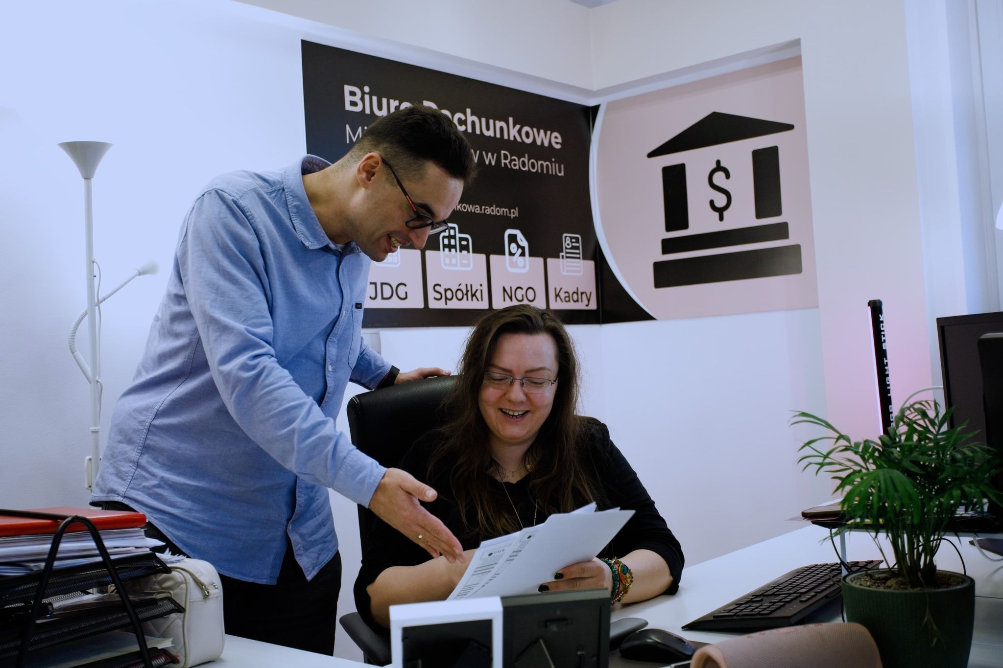 Konsultacja dokumentów w biurze rachunkowym: mężczyzna w okularach objaśnia zawartość dokumentów kobiecie przy biurku, w tle logo firmy z ikonami JDG, Spółki, NGO i Kadry.