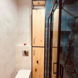 Kowadu Pracownia Sztuki Rzemiosła - Wysoka szafka łazienkowa z jasnego drewna z czarną metalową ramą, stojąca obok toalety i kabiny prysznicowej z niebieskimi płytkami.