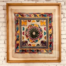 Kowadu Pracownia Sztuki Rzemiosła - Oprawiona w dębową ramę chusta z motywami słoni, lwów i kwiatów, wisząca na ścianie z białej cegły.