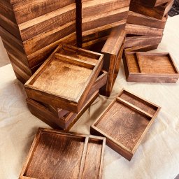 Kowadu Pracownia Sztuki Rzemiosła - Stosy brązowych, lakierowanych drewnianych pudełek i tacek, niektóre z przegródkami, ułożone na beżowej tkaninie.