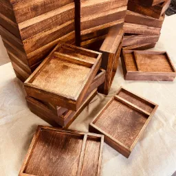 Stosy brązowych, lakierowanych drewnianych pudełek i tacek, niektóre z przegródkami, ułożone na beżowej tkaninie.