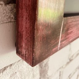 Kowadu Pracownia Sztuki Rzemiosła - Fragment drewnianej ramy w kolorze miedziano-różowym, z widoczną strukturą drewna, przylegającej do białej, chropowatej ściany.
