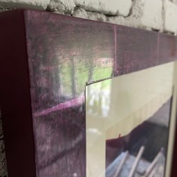 Kowadu Pracownia Sztuki Rzemiosła - Fragment drewnianej ramy obrazu w odcieniach fioletu i brązu, wiszącej na ścianie z białej cegły, z widocznym fragmentem zdjęcia wewnątrz ramy.