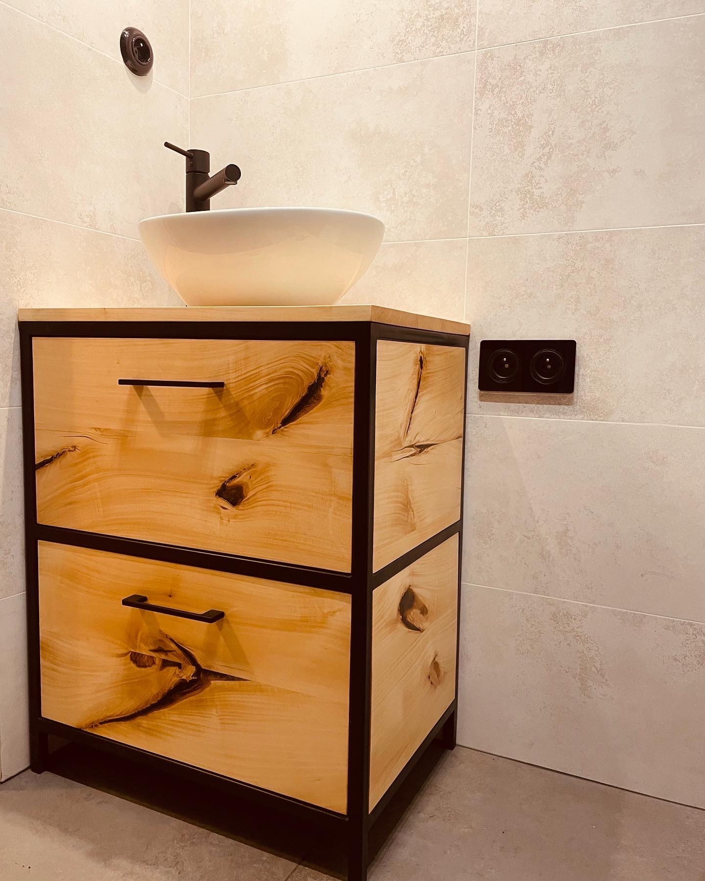 Łazienkowa szafka pod umywalkę z dwiema szufladami, wykonana z jasnego drewna z wyraźnymi słojami i czarną, metalową ramą, na której stoi biała, okrągła umywalka z czarną armaturą. W tle beżowe...