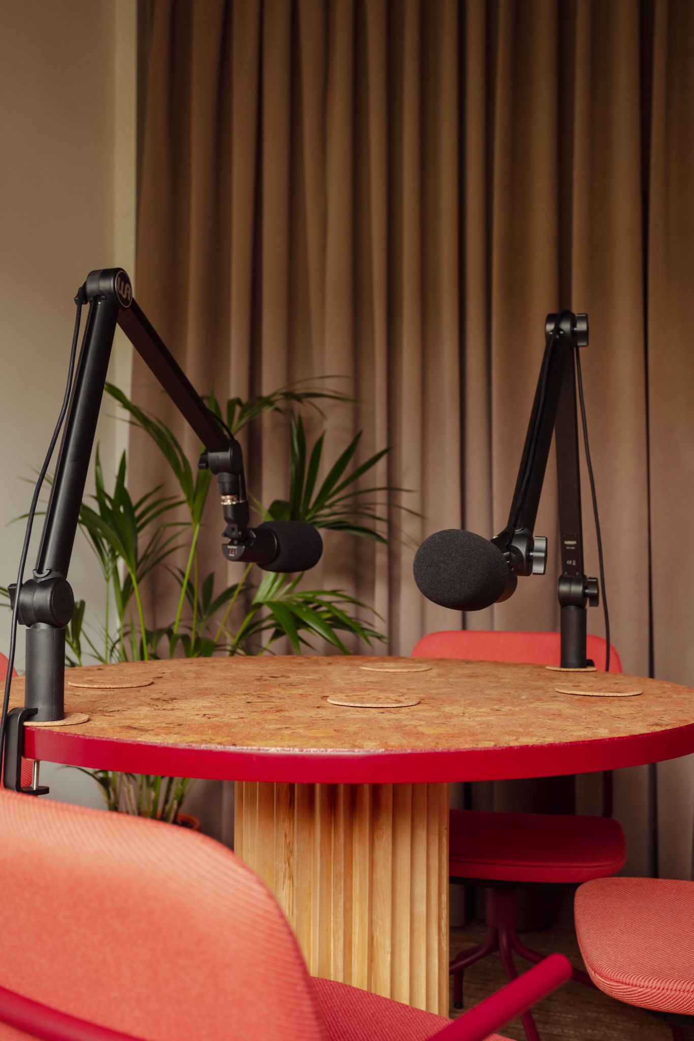 Okrągły stół z blatem z płyty OSB i czerwonym obramowaniem, otoczony czerwonymi krzesłami, z dwoma mikrofonami studyjnymi na wysięgnikach, w tle zasłony i roślina doniczkowa.
