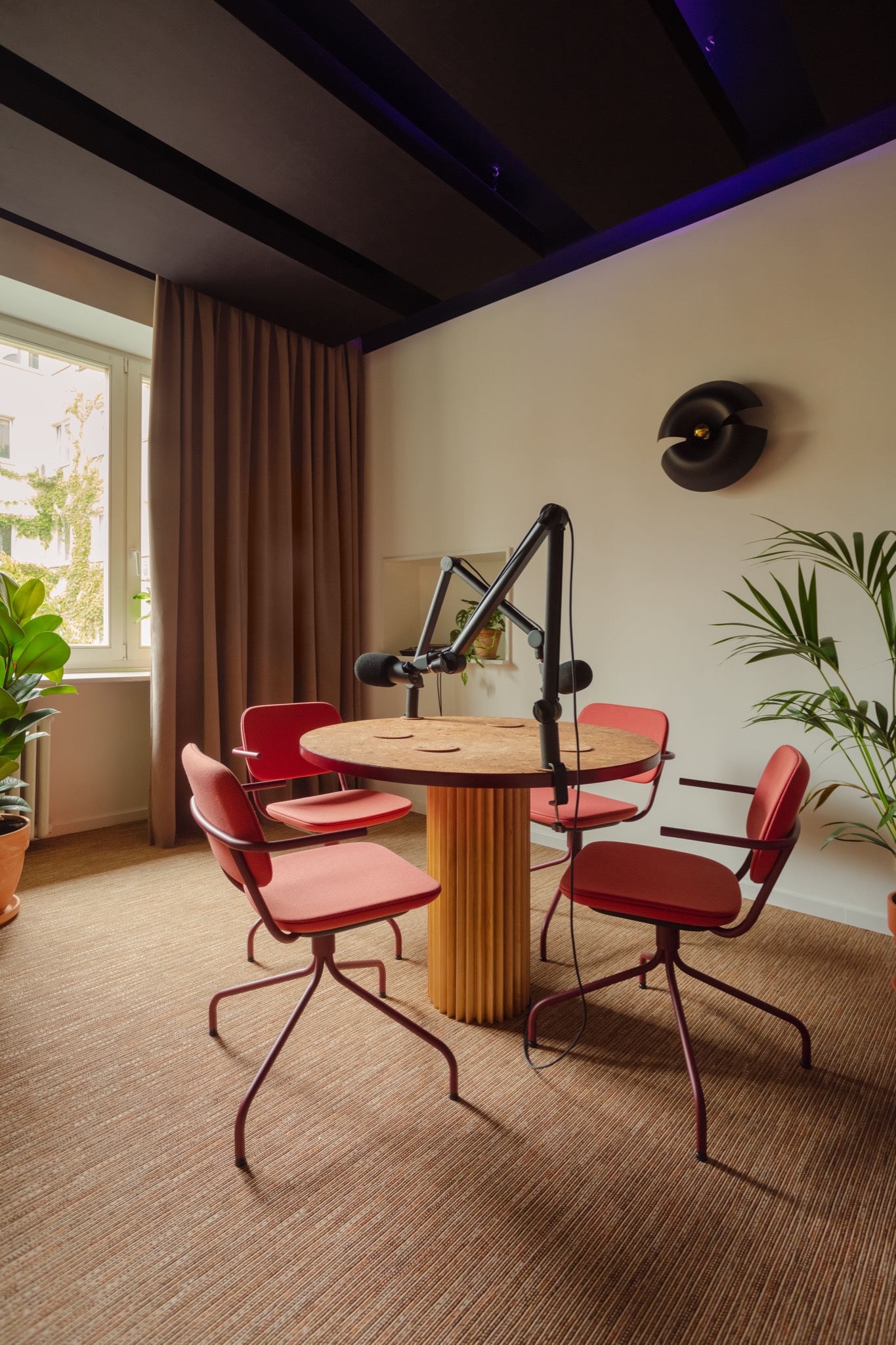 Wnętrze studia nagraniowego z okrągłym stołem z blatem z płyty OSB i podstawą z pionowych listewek, otoczonego czterema czerwonymi krzesłami na metalowych nogach, z mikrofonami na wysięgnikach...
