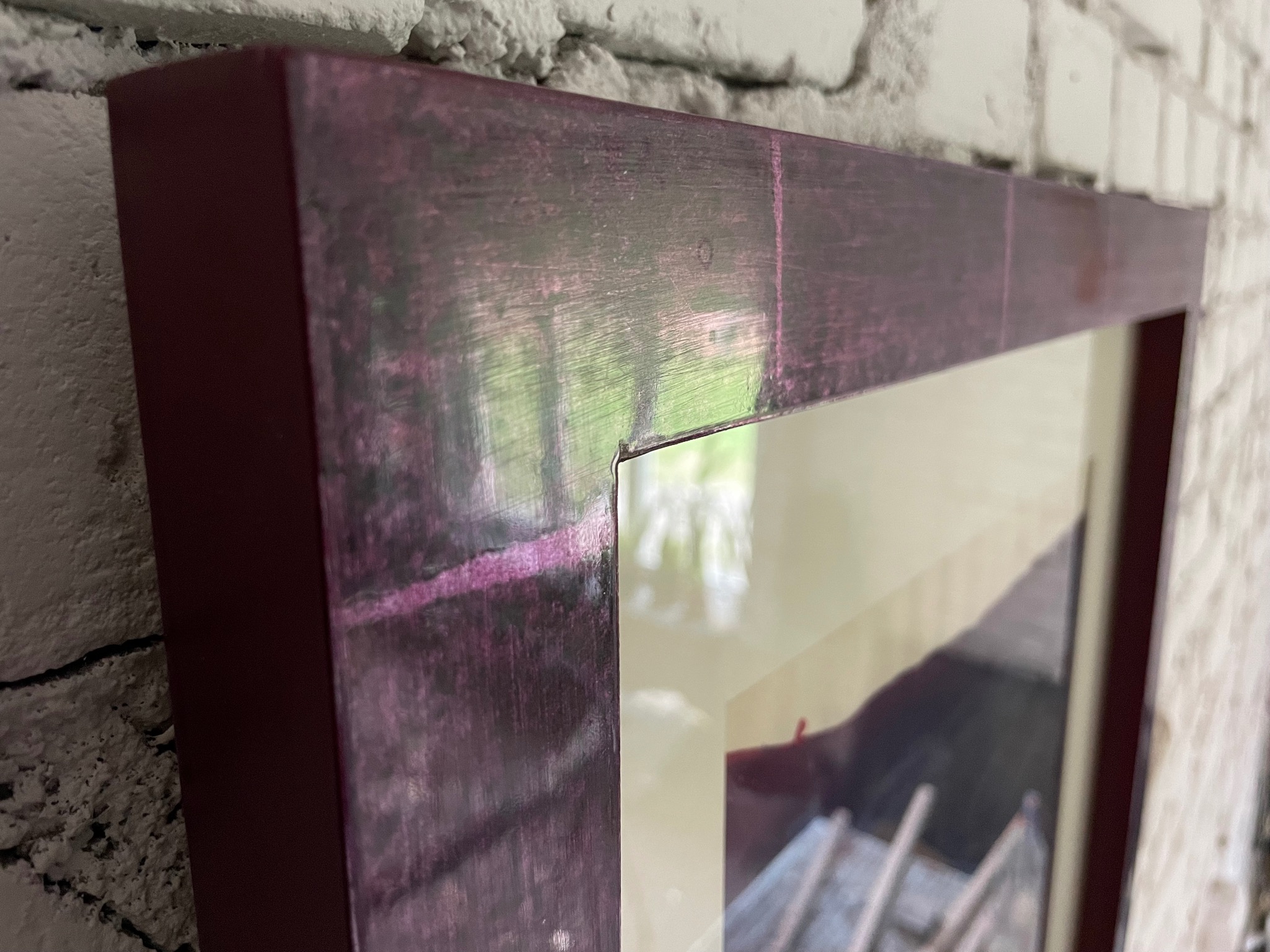 Fragment drewnianej ramy obrazu w odcieniach fioletu i brązu, wiszącej na ścianie z białej cegły, z widocznym fragmentem zdjęcia wewnątrz ramy.