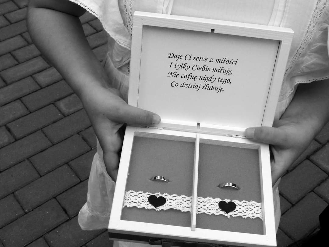 Para młoda trzyma otwarte, białe pudełko na obrączki z wygrawerowanym tekstem o miłości i przysiędze małżeńskiej; w środku dwie obrączki na koronkowej poduszeczce z czarnymi serduszkami.
