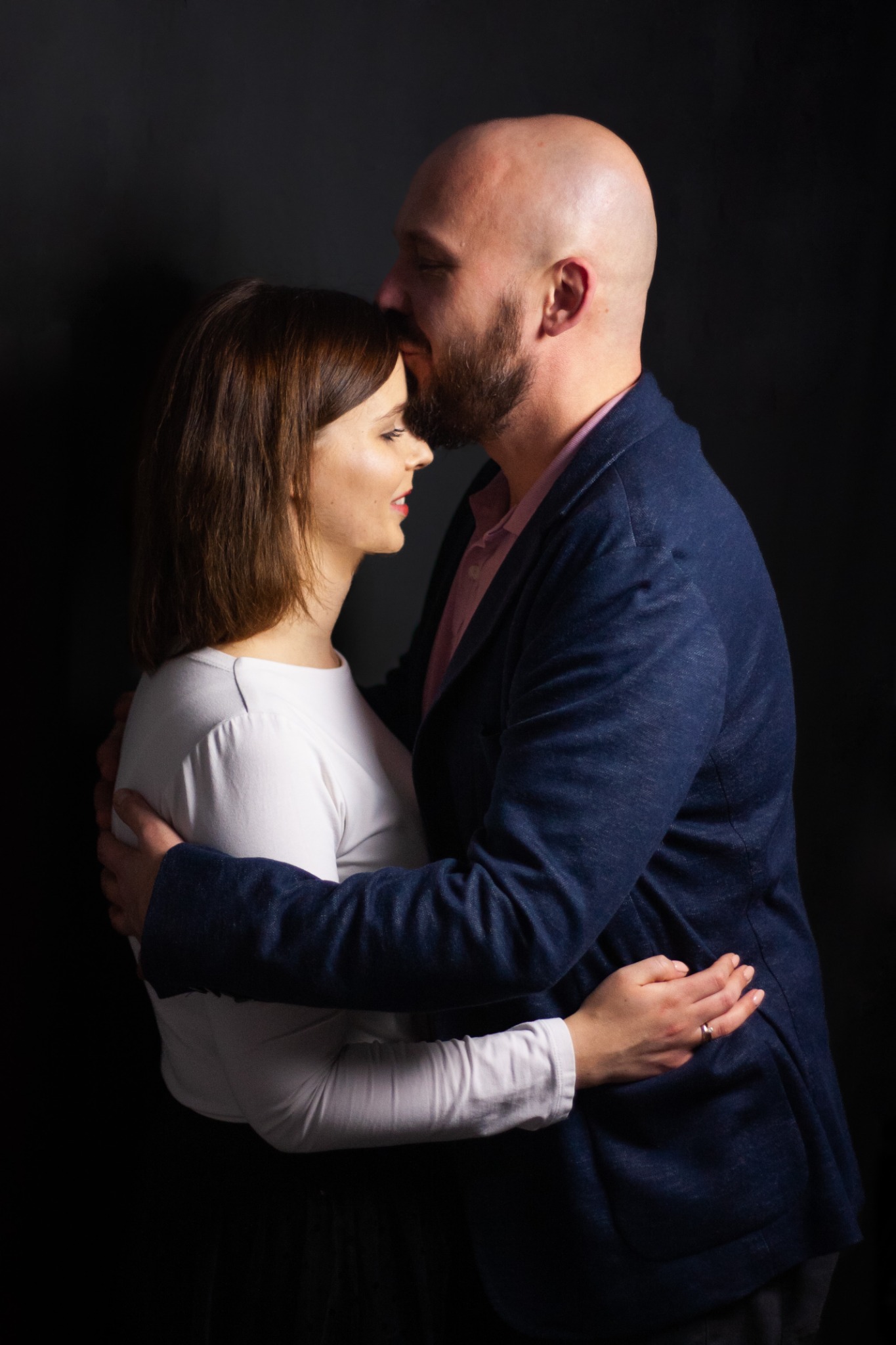 Mężczyzna w granatowej marynarce całuje kobietę w czoło na ciemnym tle, obejmując ją czule.