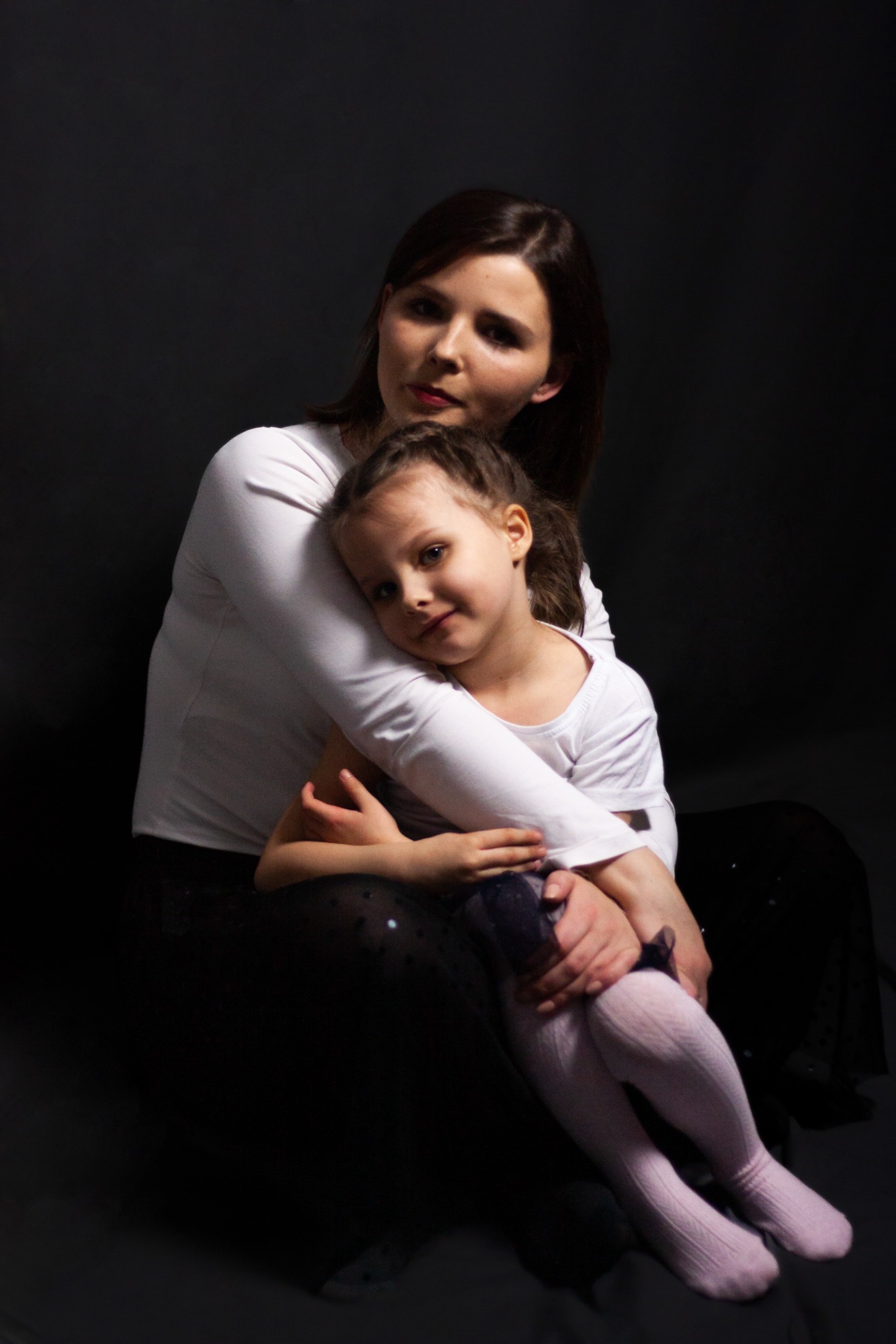 Portret matki obejmującej córkę na ciemnym tle, dziewczynka w białej bluzce i rajstopach, matka w białej bluzce z długim rękawem.