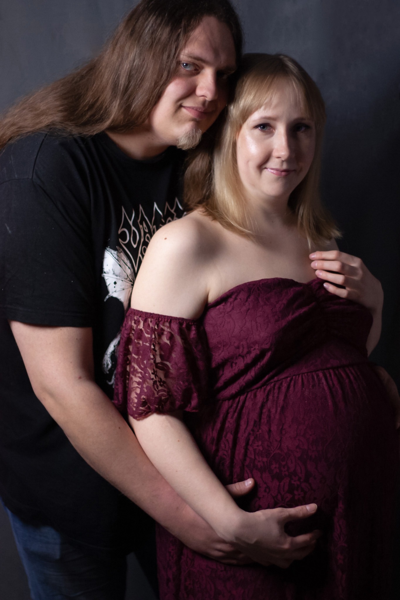 Przyszła mama w bordowej sukience z koronką, obejmowana przez partnera w czarnej koszulce z grafiką, na ciemnoszarym tle.