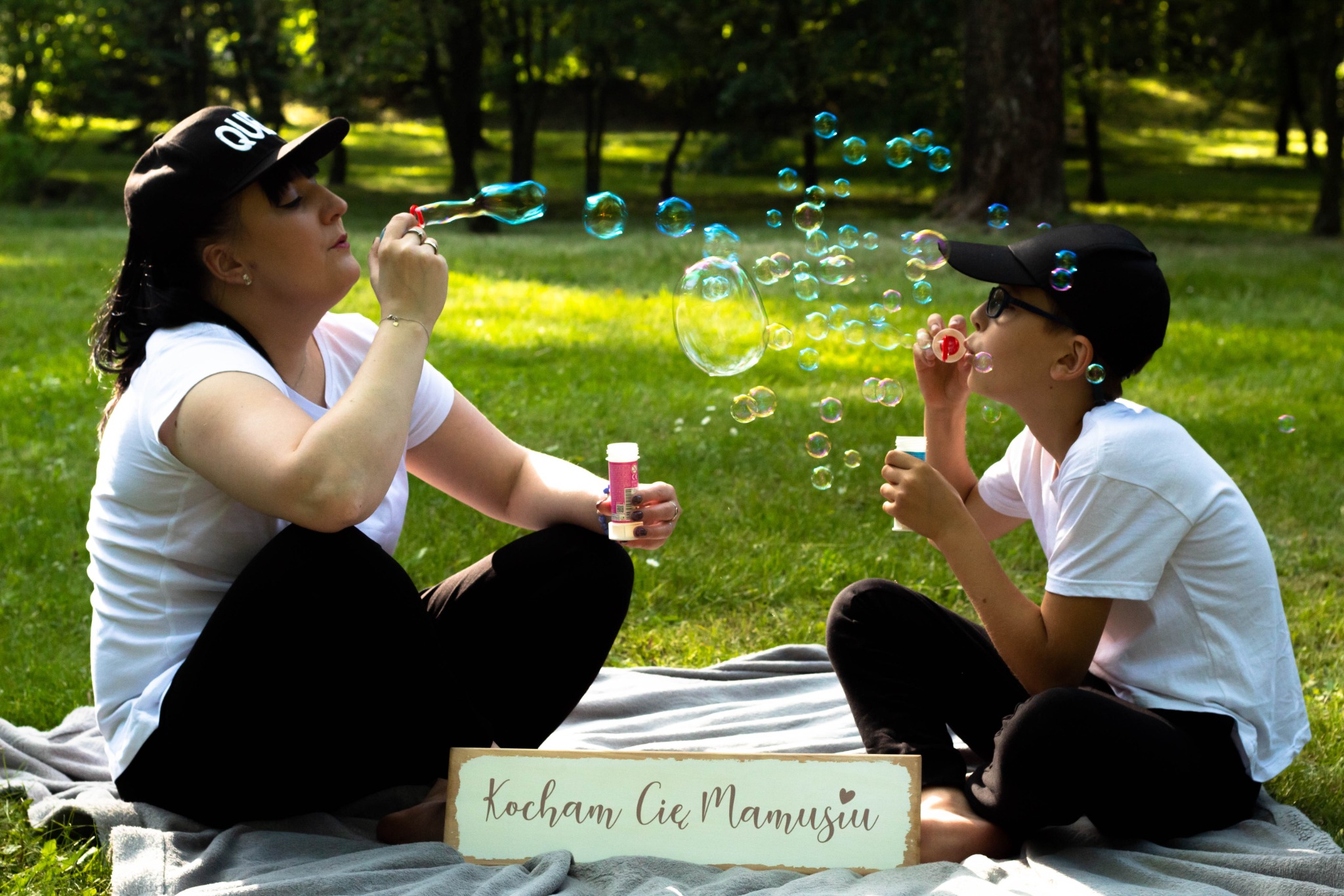 Kobieta i dziecko siedzą na kocu w parku dmuchając bańki mydlane; tabliczka z napisem 'Kocham Cię Mamusiu' leży obok.