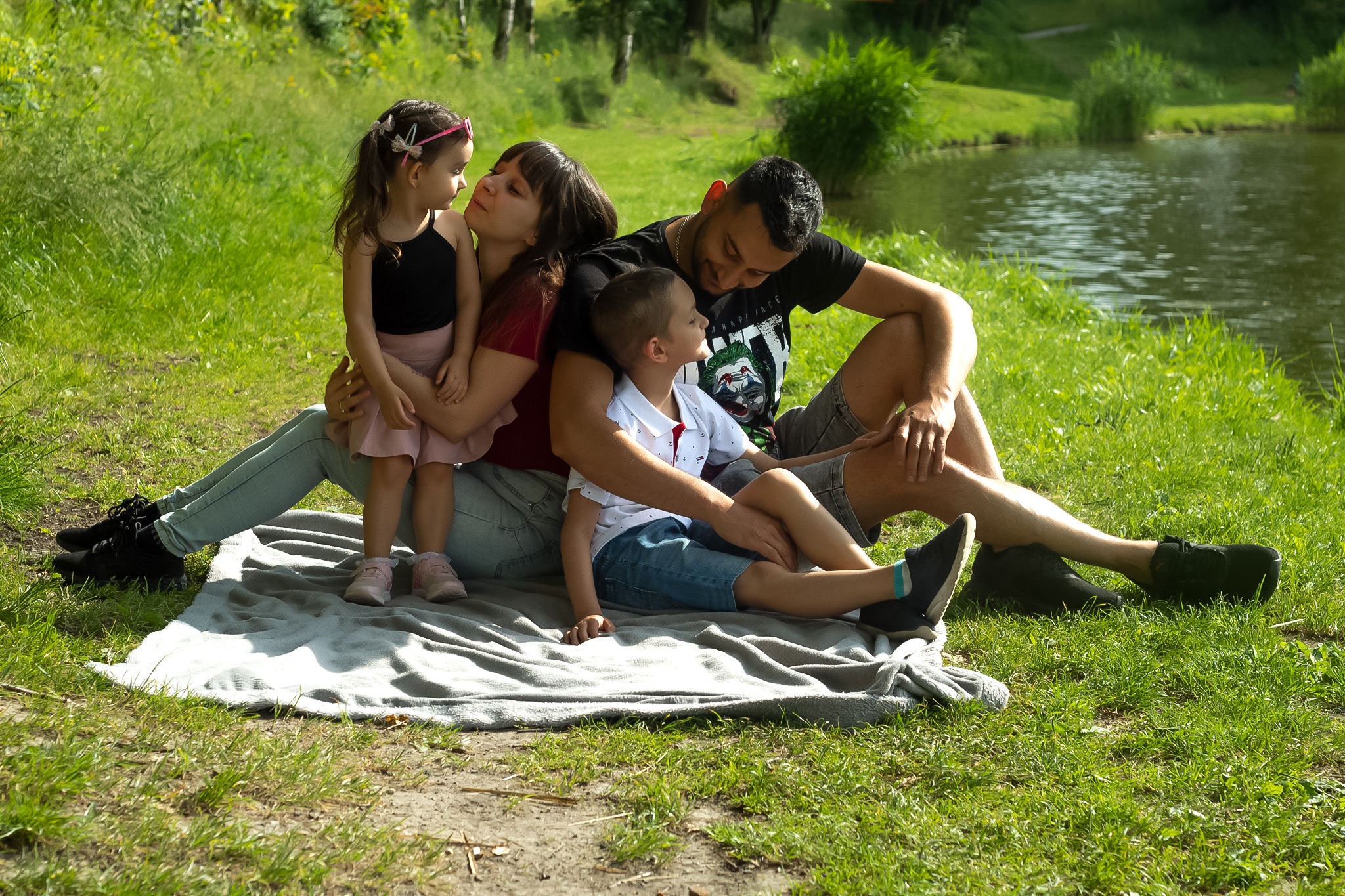 Rodzinne ujęcie w plenerze: rodzice siedzą na kocu nad brzegiem jeziora, obejmując dwójkę dzieci, mała dziewczynka z różową spinką wpatruje się w mamę, a chłopiec spogląda na tatę w koszulce...