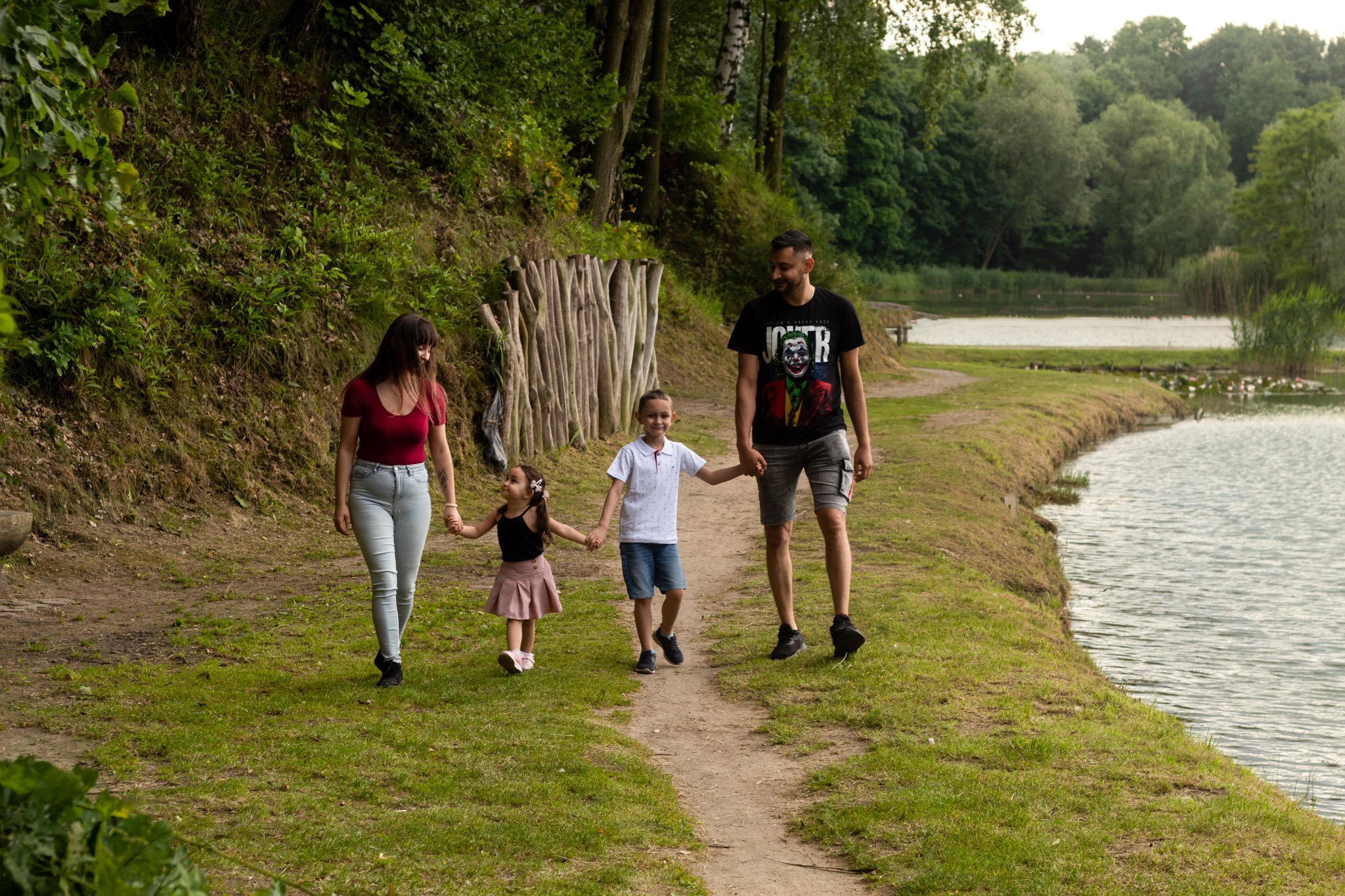 Rodzina z dwójką dzieci spaceruje ścieżką wzdłuż jeziora, tata w koszulce z Jokerem.