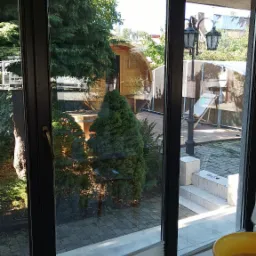 Widok przez umyte okno na zadbany ogród z drewnianą altaną, widoczny fragment żółtej miski z wodą i ślady po myciu na blacie.