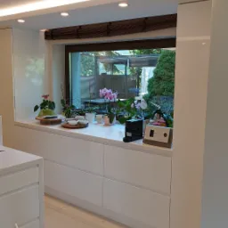 Jasne wnętrze kuchni z białymi szafkami pod oknem, na blacie doniczki z kwiatami, naczynia i sprzęt kuchenny, widok na ogród przez okno.