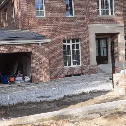 Nowo wybrukowany podjazd z kostki granitowej przed domem z czerwonej cegły z garażem, widoczne materiały budowlane i narzędzia w otwartym garażu.