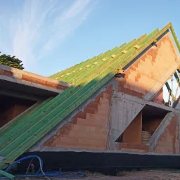 Surowy stan domu z cegły z widoczną konstrukcją dachu z zielonych łat i kontrłat, wykonany w słoneczny dzień.