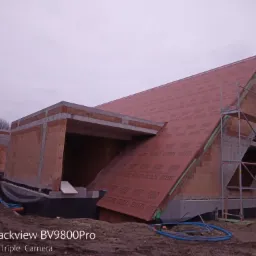 Surowy stan domu z czerwonej cegły z widocznym szkieletem dachu pokrytym czerwoną membraną, rusztowania przy szczycie dachu, niezagospodarowany teren wokół budynku.