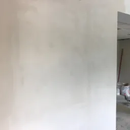 Świeżo wyszpachlowana, gładka ściana w jasnym odcieniu szarości, widoczne narzędzia malarskie i wiadro z farbą w tle, przygotowane do dalszych prac wykończeniowych w pomieszczeniu z sufitami...