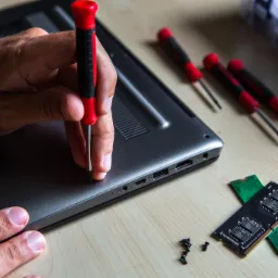 Otwieranie obudowy laptopa śrubokrętem, widoczne kości pamięci RAM i śrubki na jasnym drewnianym blacie.