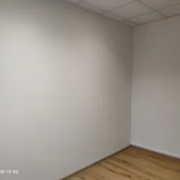 Pomalowane na jasno ściany w rogu pokoju z drewnianą podłogą, widoczne gniazdka elektryczne i sufit kasetonowy.