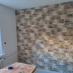 F.u.Pixel Jarosław Proszek - Ściana wewnętrzna wyłożona dekoracyjną okładziną kamienną w odcieniach beżu i szarości, widoczny fragment okna z żaluzjami, stół z niebieską taśmą malarską i fragment grzejnika.