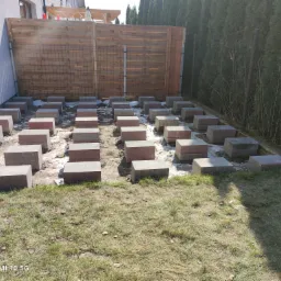 Przygotowanie podłoża pod taras: betonowe bloczki równomiernie rozmieszczone na gruncie, widoczna drewniana konstrukcja ogrodzenia z siatką, trawa i elementy izolacji.