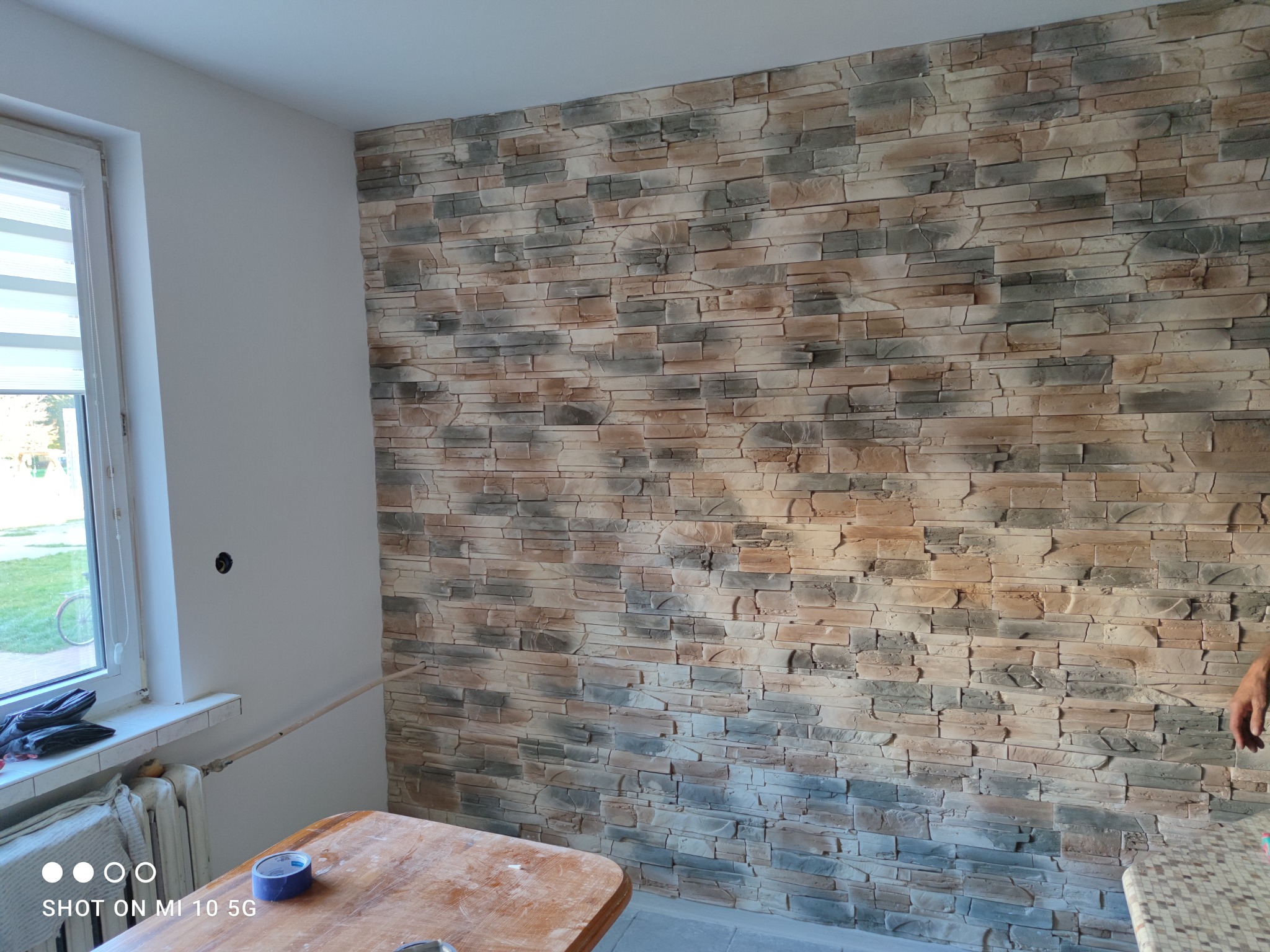 Ściana wewnętrzna wyłożona dekoracyjną okładziną kamienną w odcieniach beżu i szarości, widoczny fragment okna z żaluzjami, stół z niebieską taśmą malarską i fragment grzejnika.