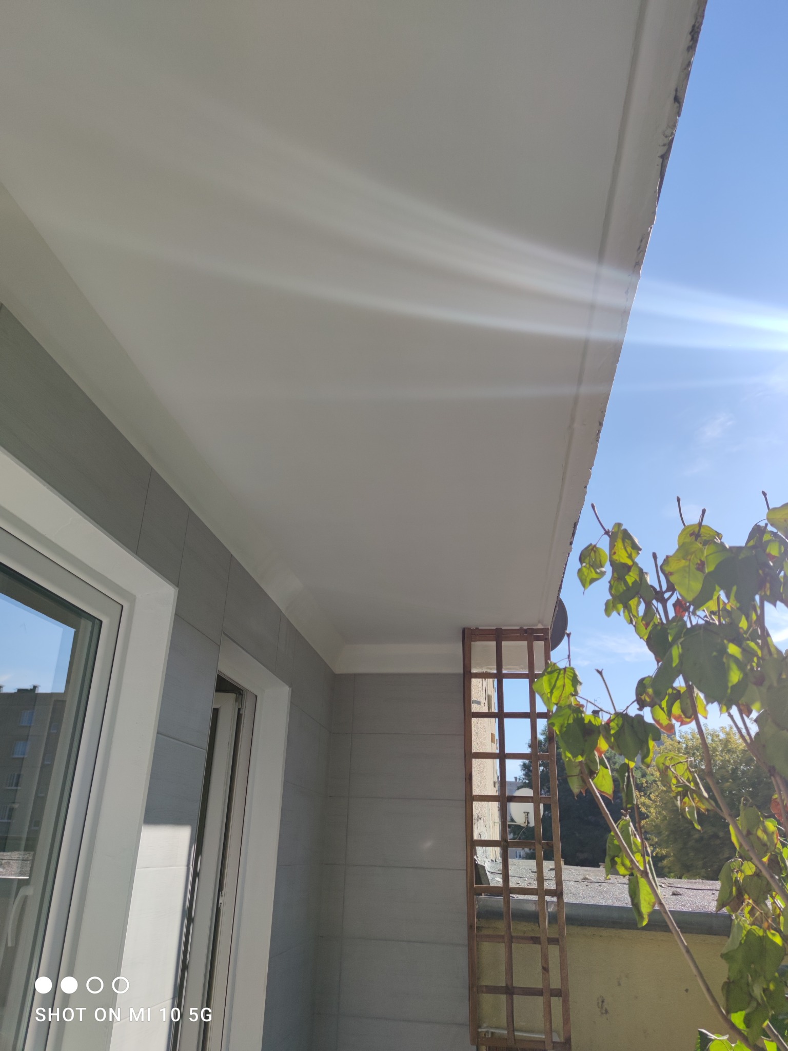 Balkon po remoncie z szarymi płytkami na ścianach, białym sufitem i oknem z widokiem na zewnątrz, z drewnianą kratką i zielonymi liśćmi w kadrze.