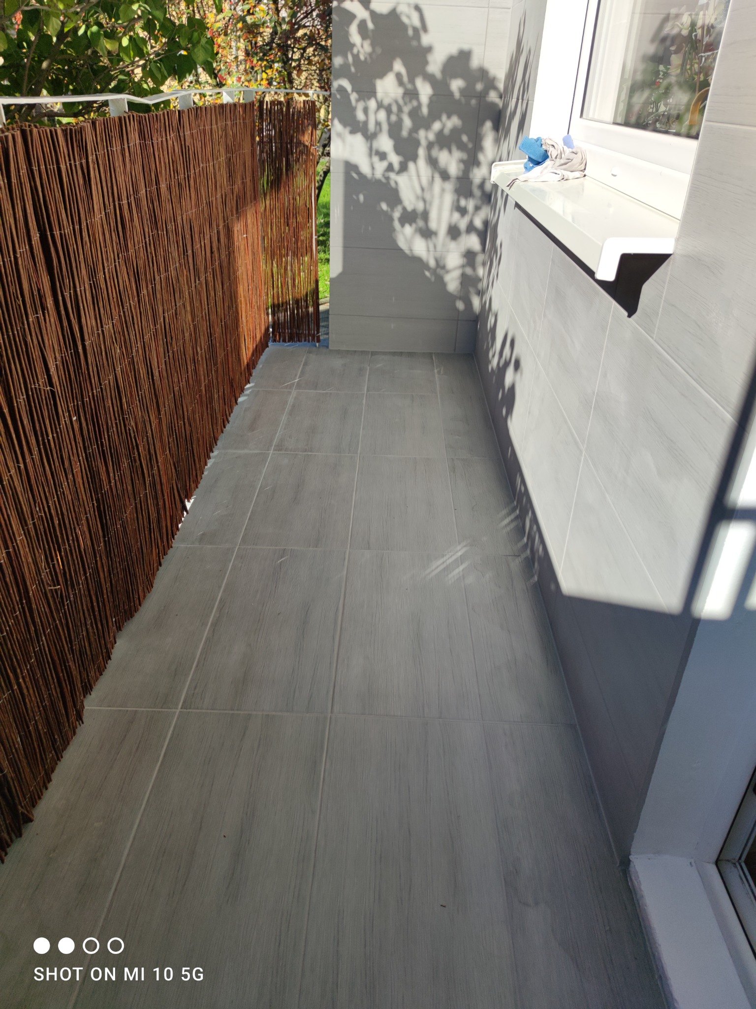 Zabudowany balkon z szarą podłogą z płytek i brązową osłoną z wikliny, widoczne cienie drzew na ścianie.