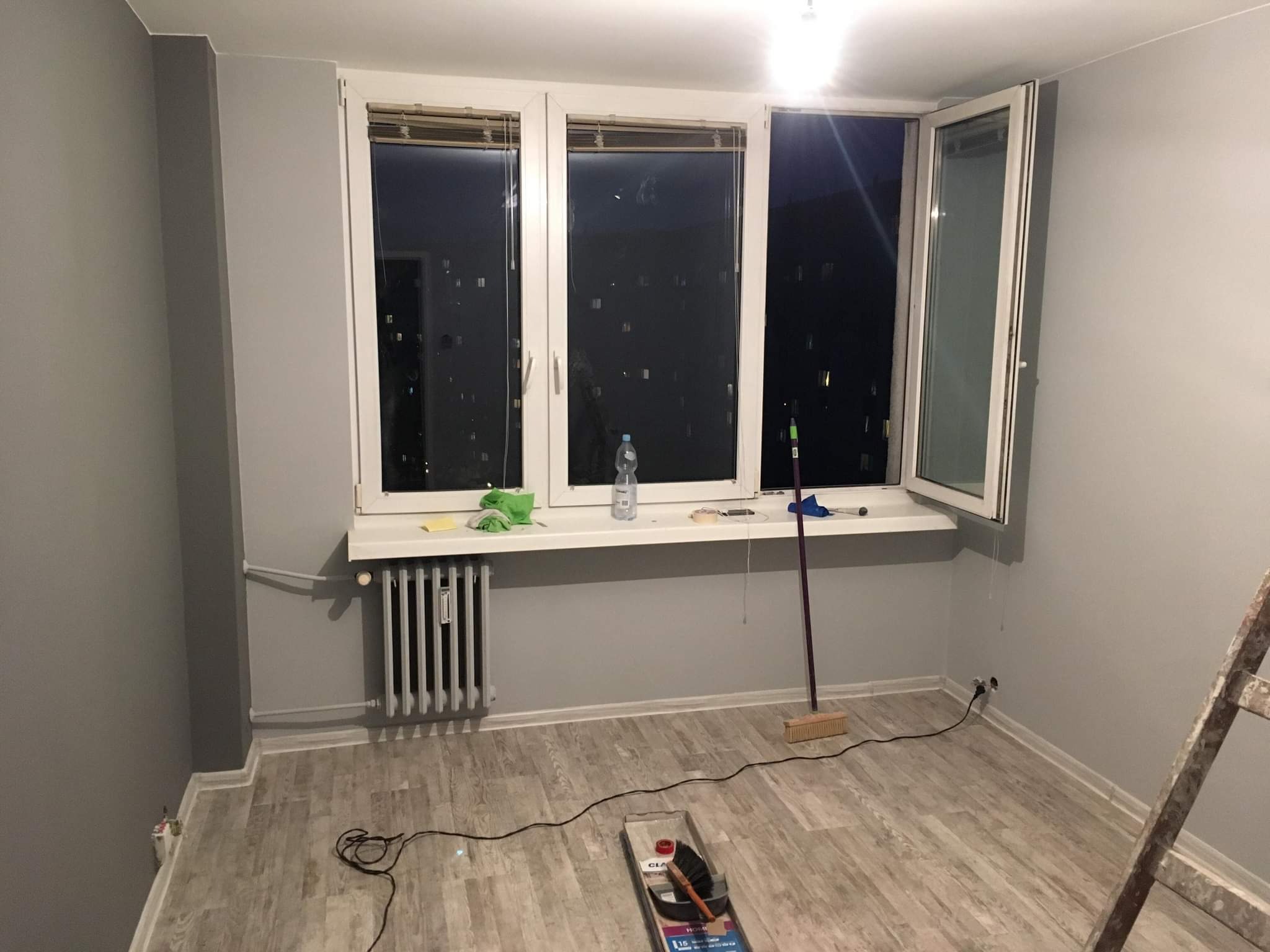 Pokój po świeżym malowaniu na szaro, widoczne narzędzia malarskie, drabina, grzejnik, okno z widokiem na nocne miasto, butelka wody na parapecie.