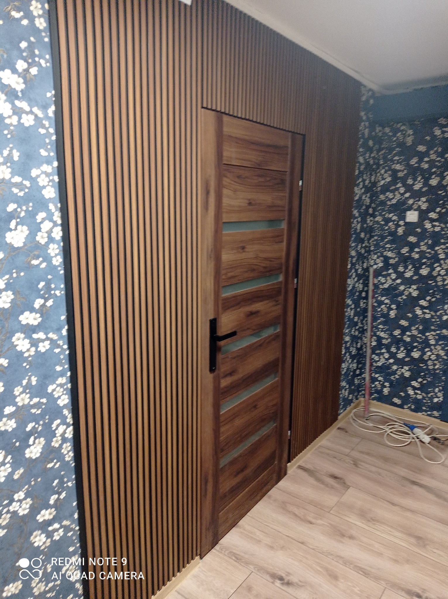 Nowoczesne wnętrze z drewnianymi lamelami ściennymi, drewnianymi drzwiami z przeszkleniami i podłogą z paneli, kontrastujące z tapetą w kwiaty na sąsiedniej ścianie.