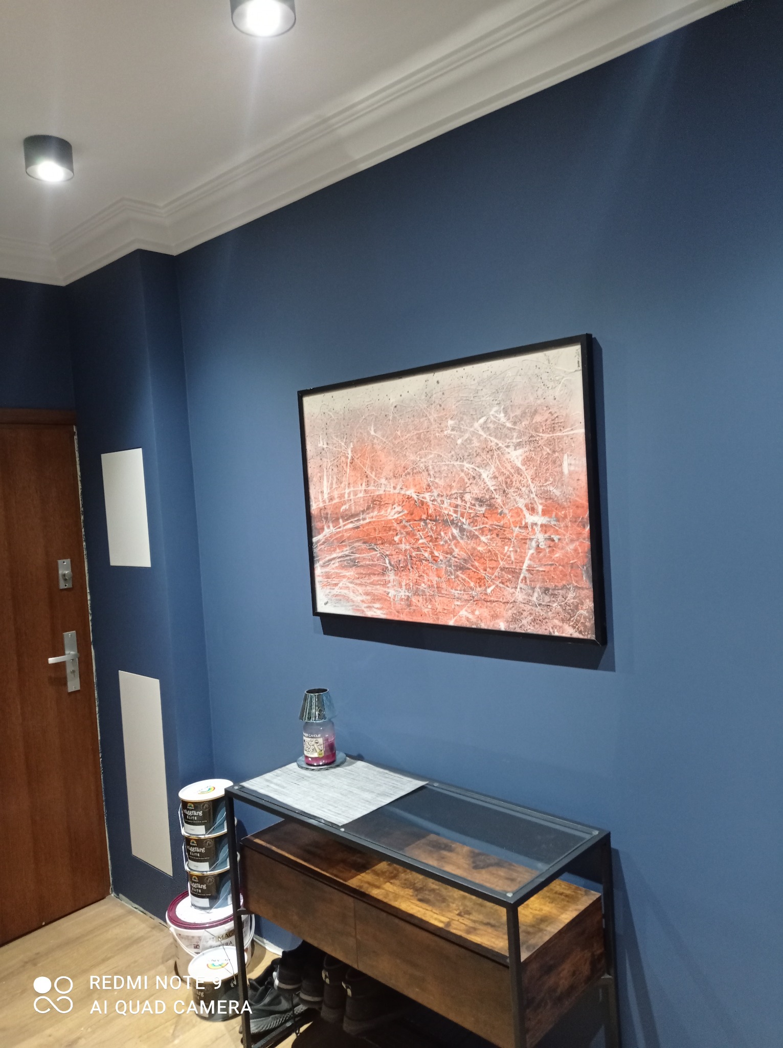 Pomieszczenie po odświeżeniu ścian farbą w odcieniach granatu, z widoczną dekoracyjną sztukaterią przysufitową, obrazem w czarnej ramie na ścianie, stolikiem z półkami oraz kilkoma puszkami farby...