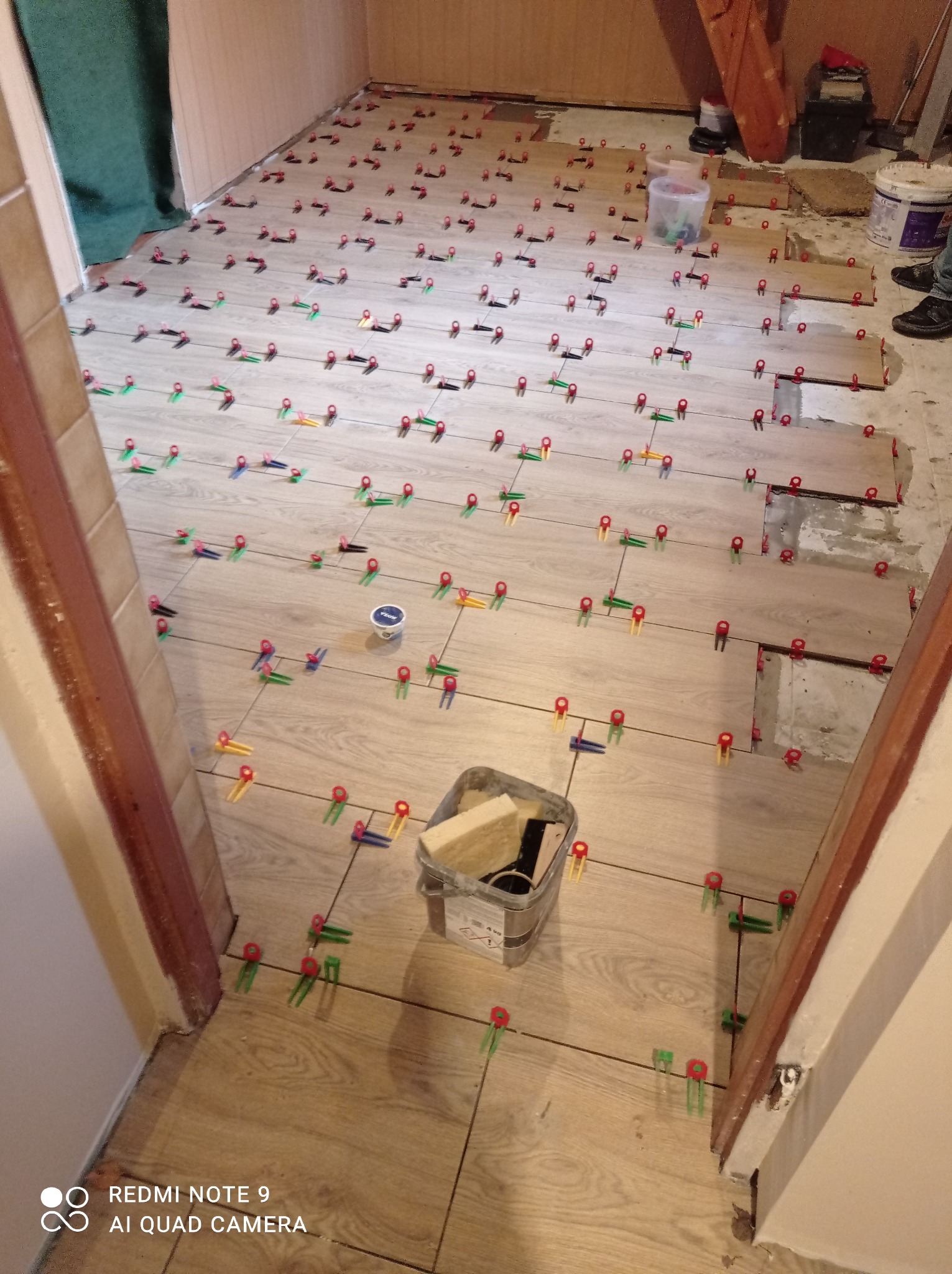 Podłoga w trakcie kładzenia płytek imitujących drewno, z systemem poziomowania płytek (klipsy i kliny) rozmieszczonym na powierzchni, widoczne narzędzia i wiadro z klejem.