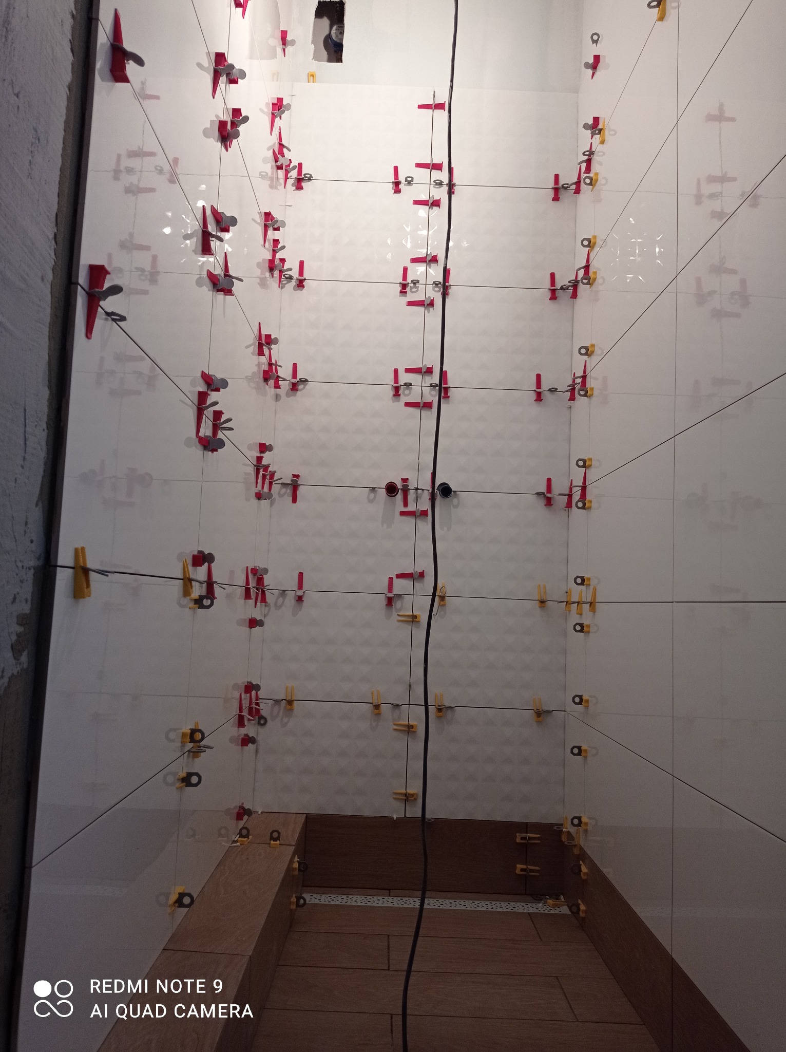 Ściana łazienki w trakcie układania białych, błyszczących płytek z użyciem systemu poziomowania (klipsy i kliny w kolorach czerwonym i żółtym), widoczny fragment brodzika wyłożonego drewnopodobną...