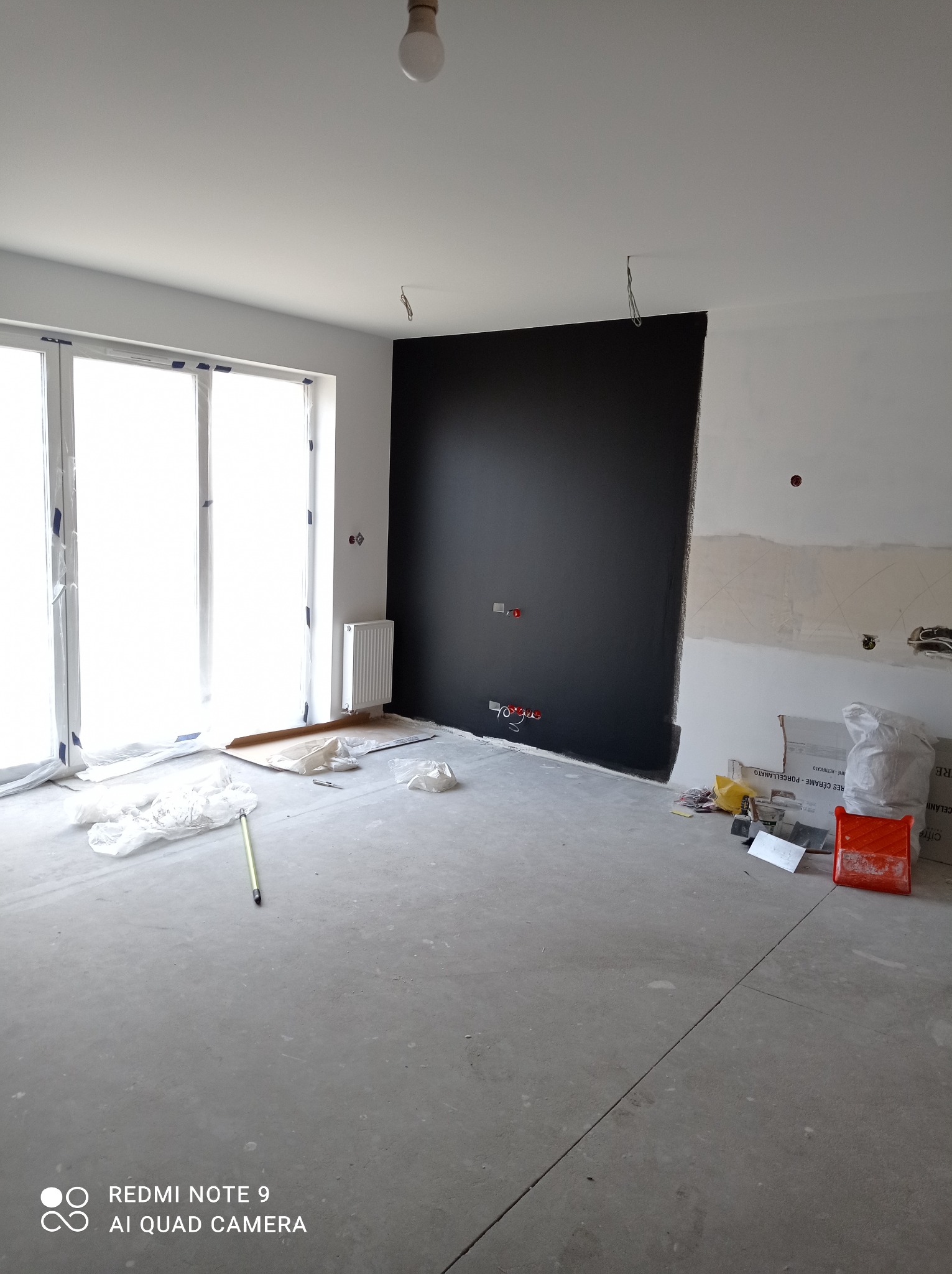Wnętrze pokoju we Wrocławiu w trakcie remontu: pomalowana na czarno ściana kontrastuje z białą, częściowo wykończoną ścianą, widoczne zabezpieczone folią okno balkonowe, grzejnik, narzędzia...