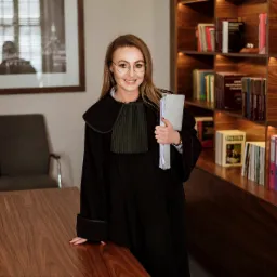 Uśmiechnięta adwokat w todze, trzymająca dokumenty, na tle biura z drewnianym biurkiem i regałem pełnym książek prawniczych.