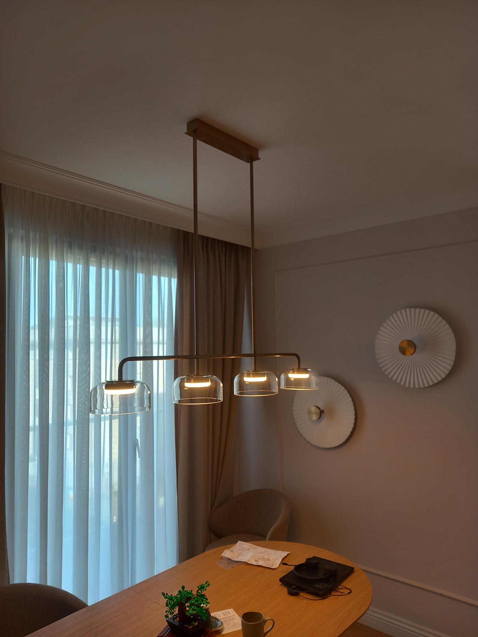 Nowoczesna lampa wisząca z czterema szklanymi kloszami, oświetlająca drewniany stół z dekoracjami. W tle okno z zasłonami i dekoracyjne talerze na ścianie.