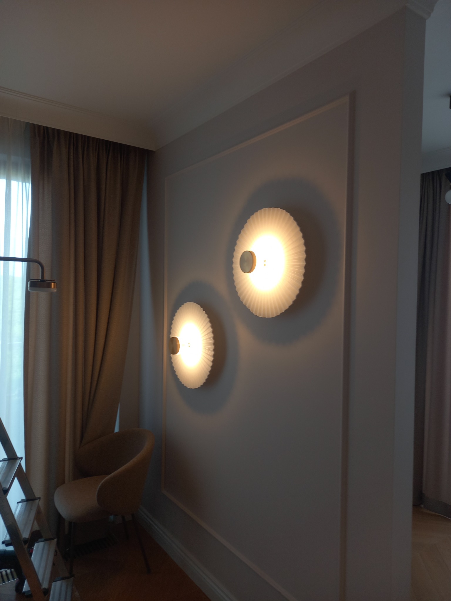 Dwa okrągłe, dekoracyjne kinkiety z żarówkami LED, zamontowane na szarej ścianie z ozdobnymi listwami. W tle zasłony i krzesło, widoczny fragment drabiny.
