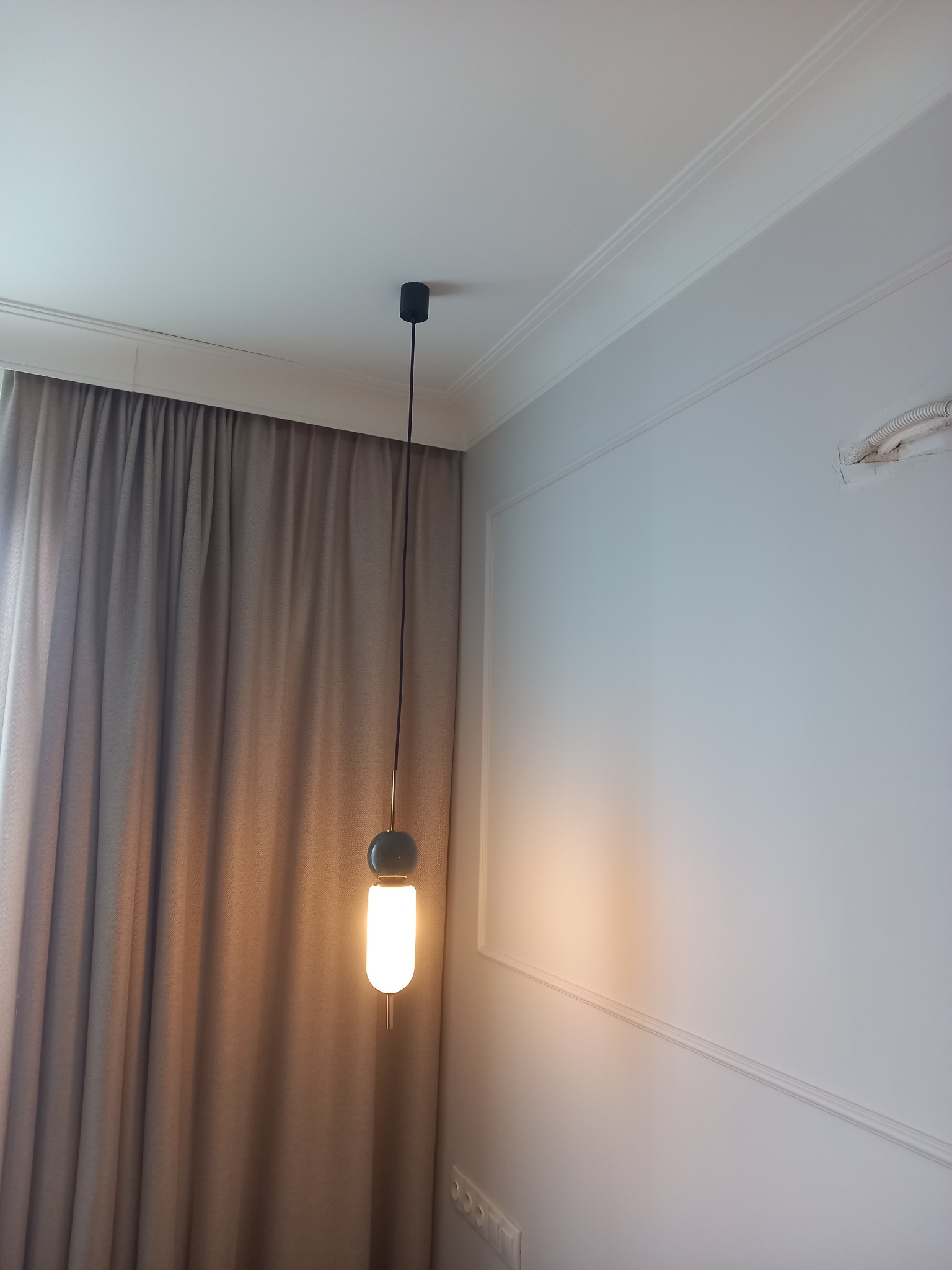 Nowoczesna lampa wisząca z ciepłym światłem, zwisająca na tle zasłon i białej ściany z dekoracyjną listwą. Widoczne gniazdka elektryczne i fragment instalacji.