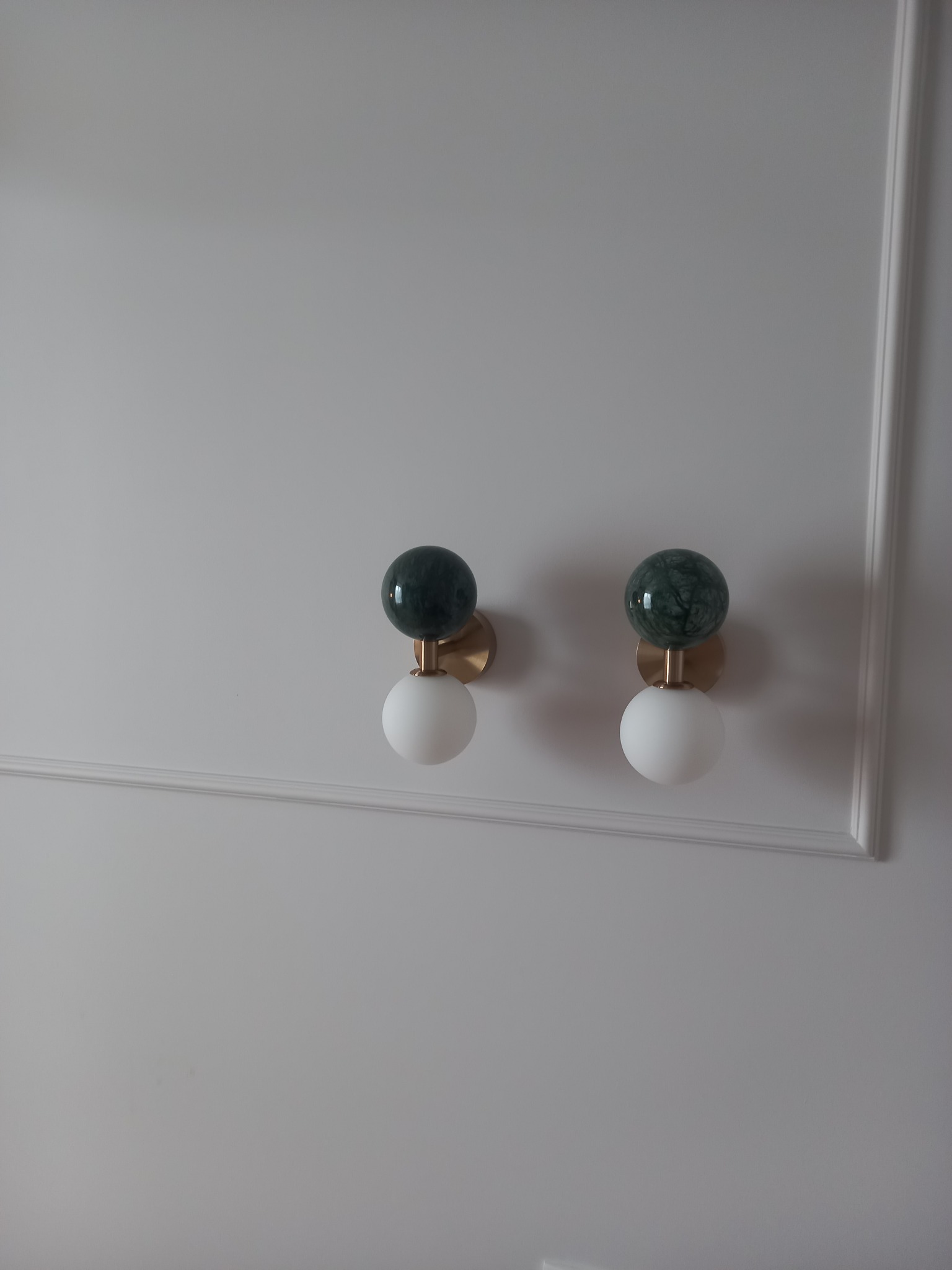 Dwa nowoczesne kinkiety kuliste z zielonym marmurem i białym szkłem, zamontowane na białej ścianie z dekoracyjną listwą. Minimalistyczny design oświetlenia.