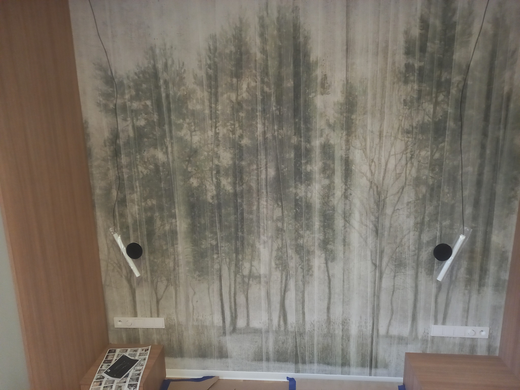 Pokój z fototapetą leśną i zamontowanymi lampami wiszącymi. Widoczne gniazdka elektryczne i fragmenty mebli. Oświetlenie w trakcie montażu.