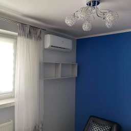Eko-Klima Krzysztof Żochowski - Biały klimatyzator zamontowany na szarej ścianie nad półkami w pokoju z niebieską ścianą, obok okna z firanką i pod ozdobnym żyrandolem.